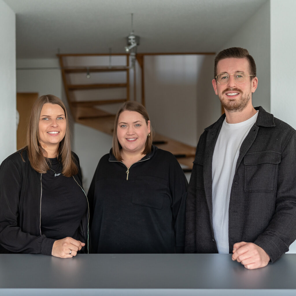 Drei Mitarbeitende von Steuerberater Ellermann, Jungmann und Klippert in Luedenscheid stehen gemeinsam im modernen Buero und laecheln in die Kamera.